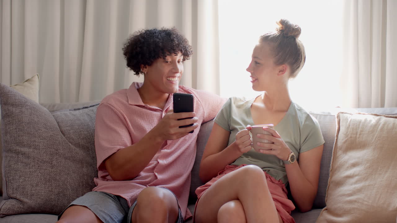 Smiling diverse couple relaxing on couch, enjoying coffee and smartphone together, at home