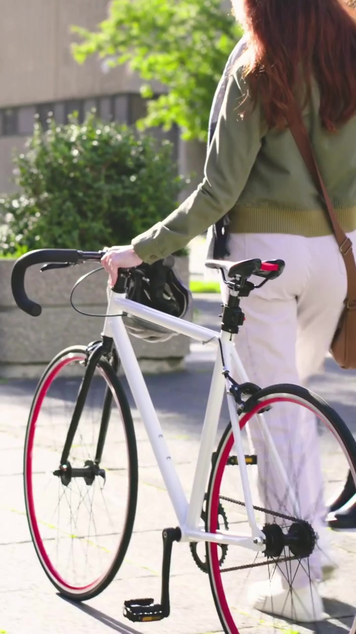 Vertical video: Walking woman in green jacket pushing white bike into shaded path, helmet and bag