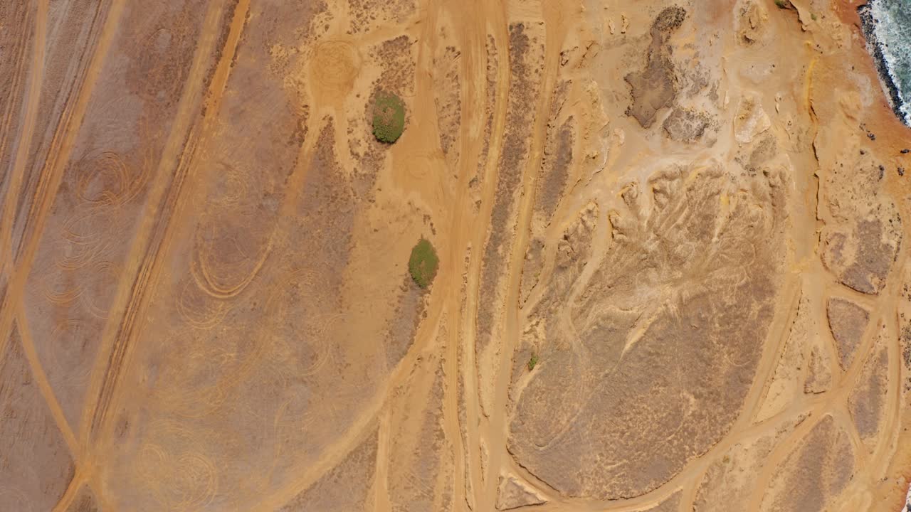 From a vertical drone angle, intersecting tire loops lace an ochre desert plain, forming swirls and arcs across packed sand, a graphic tangle of motion and human traces