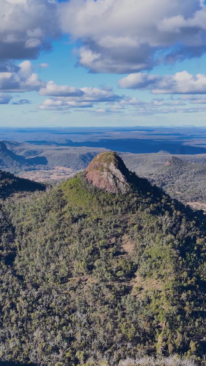 La telecamera di un drone panoramica fluidamente attraverso le cime e le valli boscose del Parco Nazionale Warrumbungle, rivelando un terreno accidentato, nuvole spettacolari e orizzonti lontani alla luce del giorno.