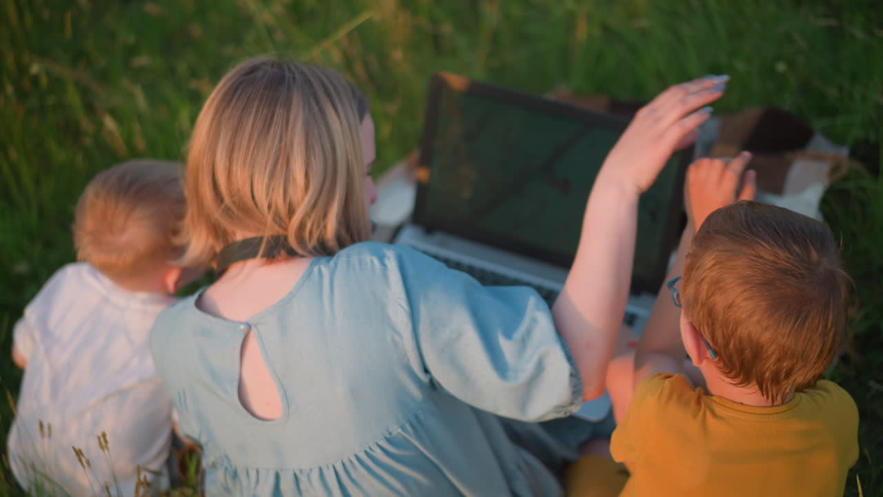 노란색 옷을 입은 어머니가 노트북을 들고 잔디에 앉아있는 뒤쪽 사진. 노란색을 입은 그녀의 어린 아들은 그녀와 장난스럽게 상호 작용하는 반면, 색으로 입은 다른 아들은 그녀의 에 조용히 앉아 있습니다.