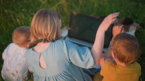back shot of a mother in a blue gown sitting on a grassy field with a laptop. her young son in yellow playfully interacts with her, while her other son, dressed in white, sits quietly by her side