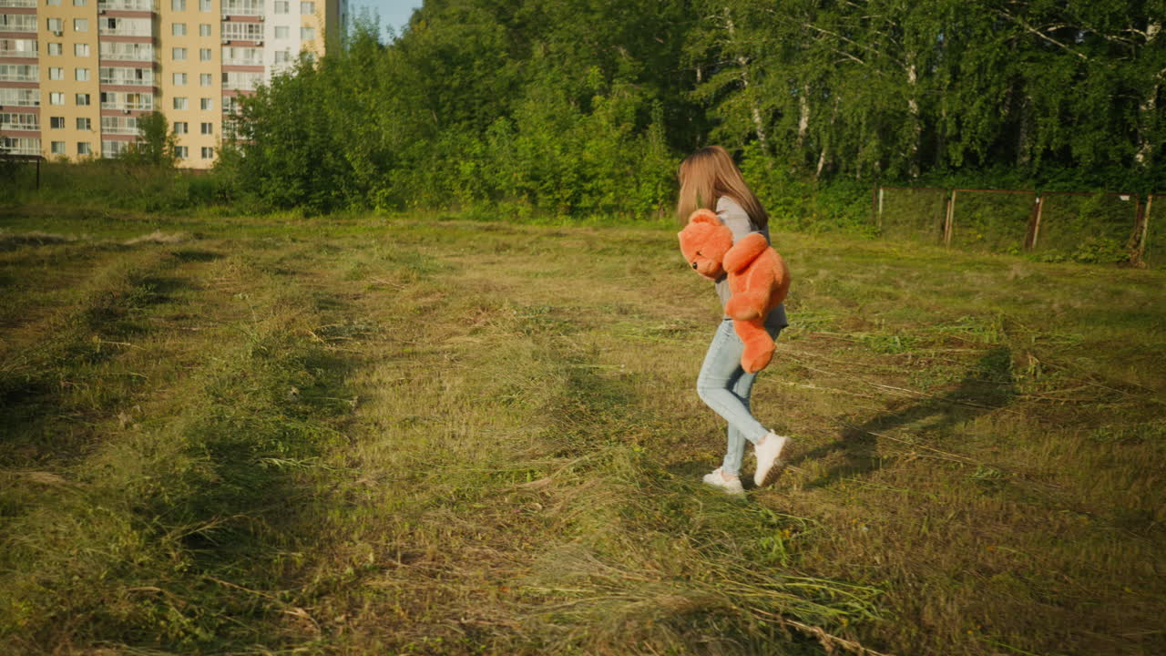 Side view of young woman walking across grassy farmland holding teddy bear under arm, with tall apartment buildings in background and green trees surrounding open area on sunny day