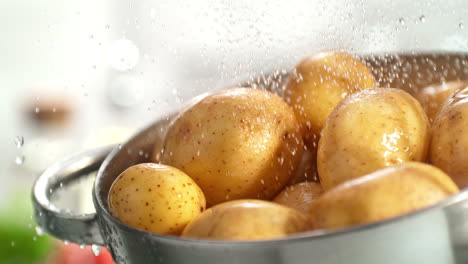 patatas sin pelar temprano enjuagadas en un filtro bajo gotas de agua salpicando en cámara lenta en una cocina brillante