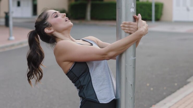 Gripping metal pole, adult woman stretching back on quiet street for exercise, in workout clothes