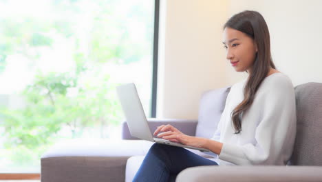 close up of a young woman sitting on a couch typing on the laptop balanced on her legs
