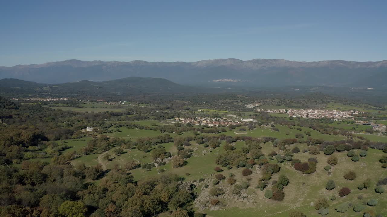 vuelo lateral en un valle con el sistema montañoso central de la península ibérica en el fondo donde hay una niebla llamativa vemos un centro urbano rural y colinas con una variedad de arbustos