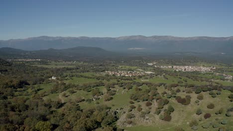 Vuelo-Lateral-En-Un-Valle-Con-El-Sistema-Montañoso-Central-De-La-Península-Ibérica-Al-Fondo-Donde-Hay-Una-Llamativa-Neblina-Vemos-Un-Centro-Urbano-Rural-Y-Cerros-Con-Variedad-De-Arbustos