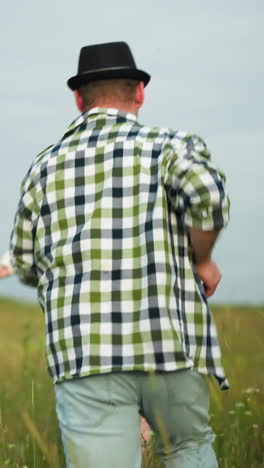 a man in a black hat and checkered shirt runs after a woman in a white dress and hat through a grassy field under a cloudy sky. the scene captures a dynamic and playful moment in a rural landscape