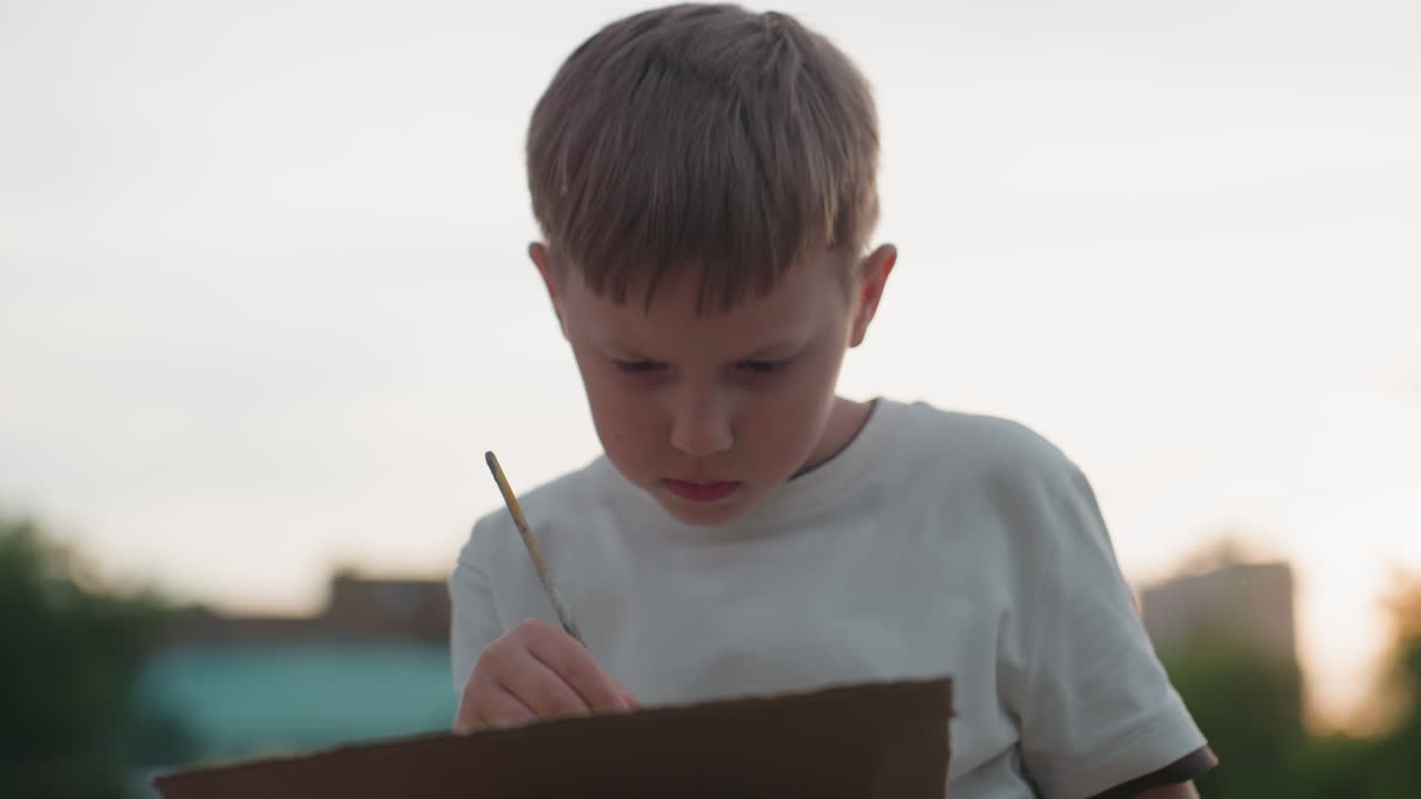 young schoolboy seated on wooden dock under soft sunset light, concentrated on painting with fine brush on board, applying precise strokes with focused expression during warm evening art practice
