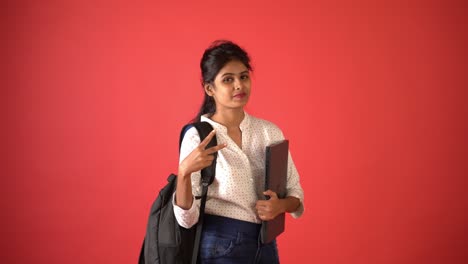 Una-Joven-India-Con-Pantalones-Blanco,-Portátil-Y-Mochila-Escolar-Con-Símbolo-De-Paz-En-La-Mano-Parada-En-Un-Fondo-Rojo-Aislado