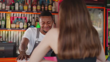 positive barman listens to long-haired woman in bar. smiling black bartender accepts order slowly wiping countertop. pleasant interaction with customer