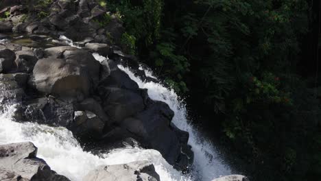 a bright cascade of clear water tumbles forcefully over dark volcanic boulders, disappearing into a dense green forest, highlighting the wild beauty of an untouched natural landscape.