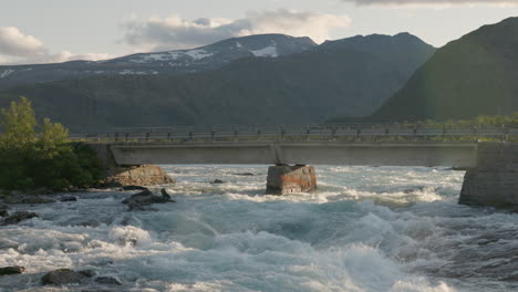 Una-Furgoneta-Azul-Que-Circula-Por-Un-Hermoso-Puente-En-Las-Montañas-Noruegas,-Rodeada-De-Un-Hermoso-Paisaje-Y-Un-Río-De-Aguas-Bravas.