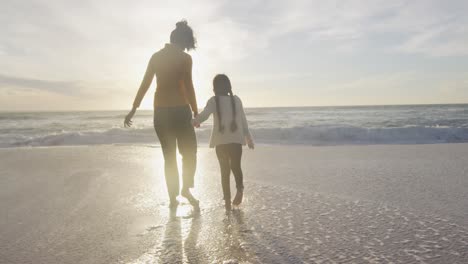 Back-view-of-hispanic-mother-and-daughter-walking-on-beach-at-sunset