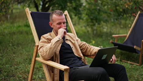 happy man working remotely in the garden having online chat on his computer. freelance worker talking to clients online sitting in the nature.