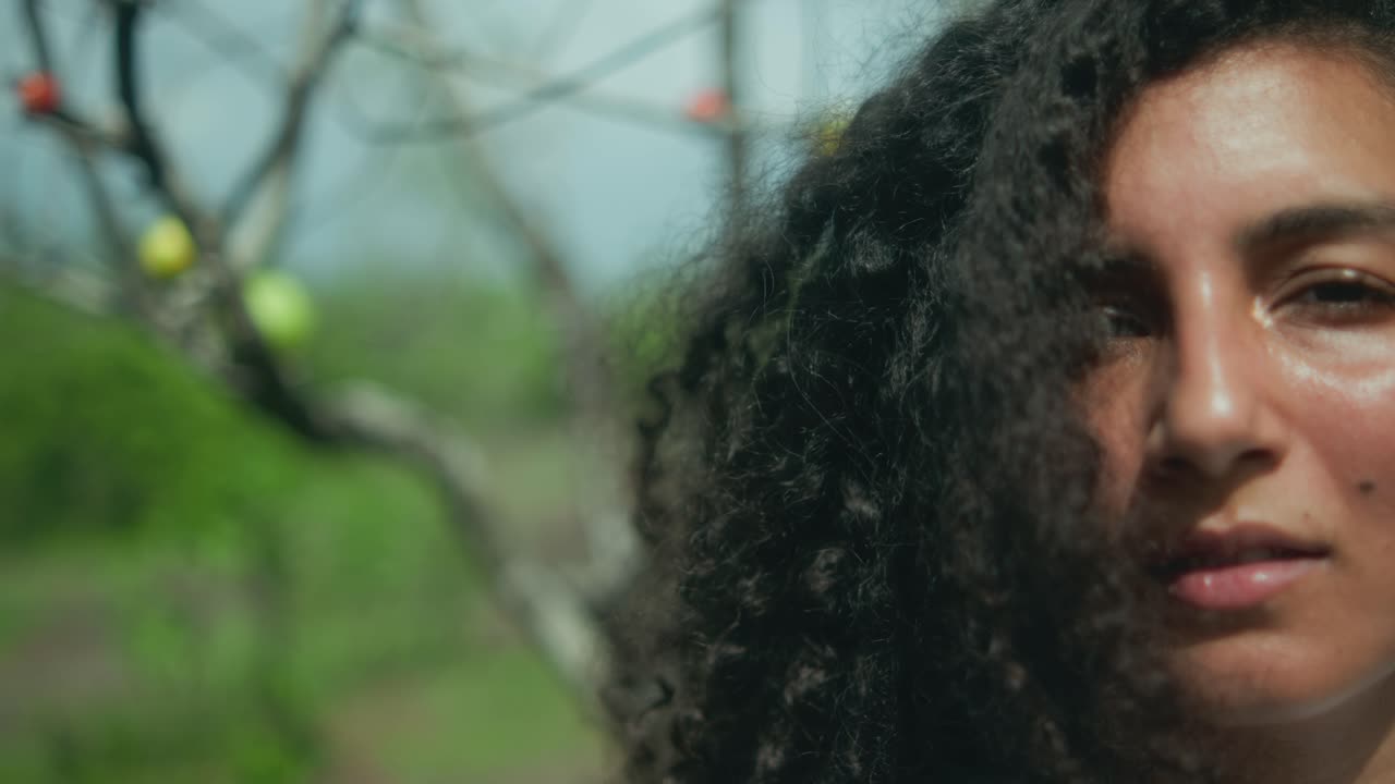 Extreme close-up of half a woman’s face with loose curly hair, with a tree and vegetation in the blurred background