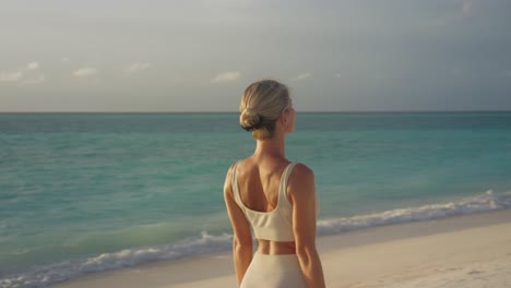 Detrás-De-Una-Atractiva-Mujer-Rubia-Estirando-Los-Brazos-Hacia-Arriba-Haciendo-Yoga-En-Una-Playa-Tropical