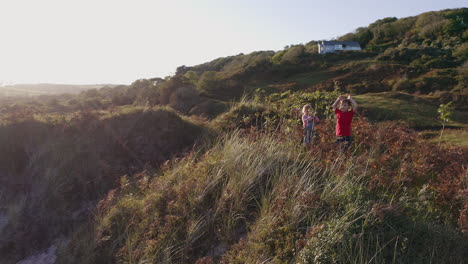 Toma-De-Drone-De-Un-Niño-Y-Una-Niña-En-Vacaciones-En-La-Playa-Saludando-Mientras-Juegan-En-Dunas-De-Arena-Con-Luz-Nocturna