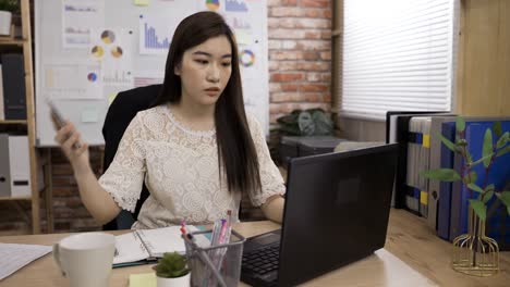 asian woman secretary receiving urgent call from her boss while working at desk is picking up her knitted jacket and hurrying out of the office during daytime.