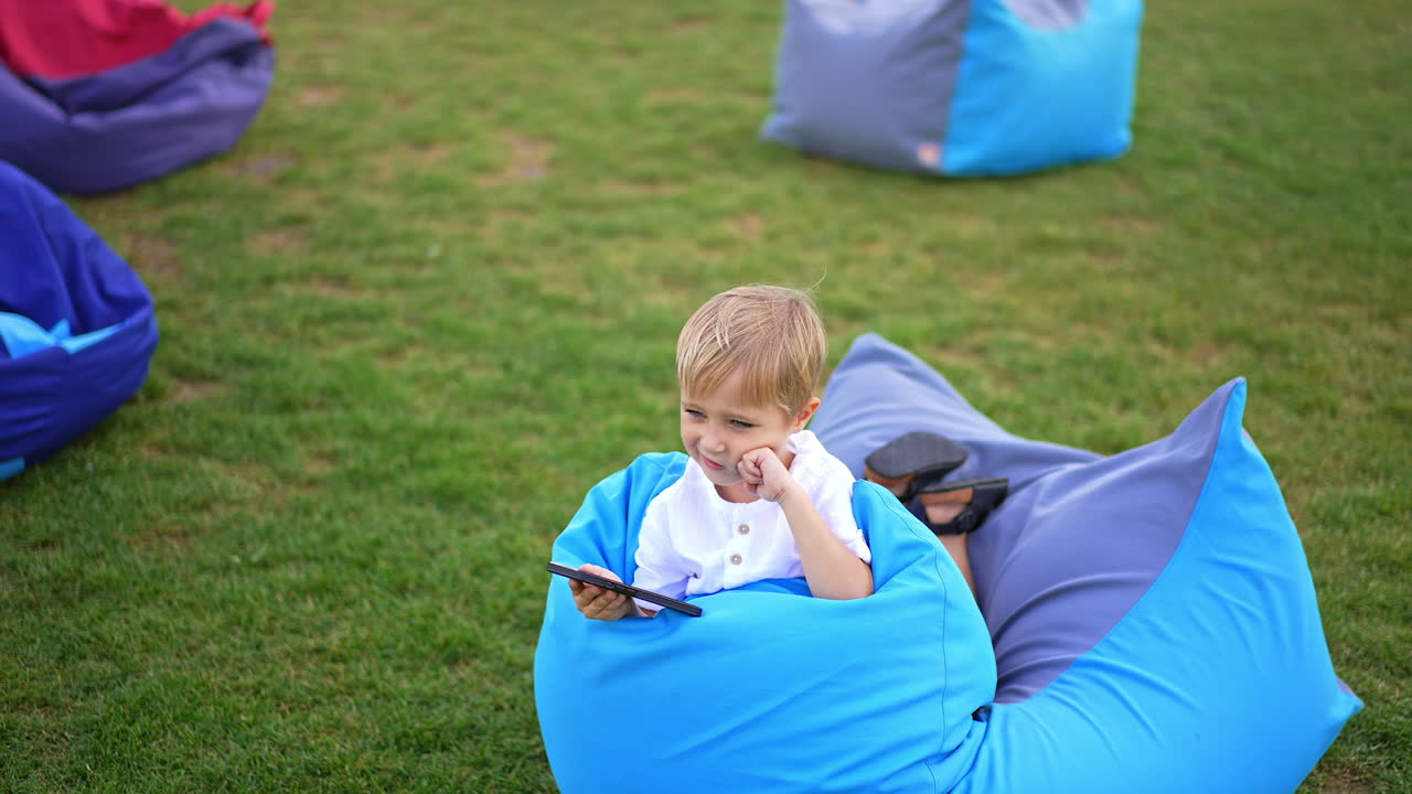 Blond preschooler having rest in bean bag chair outside. Cute boy holding phone in hand but looks aside. Circle footage.
