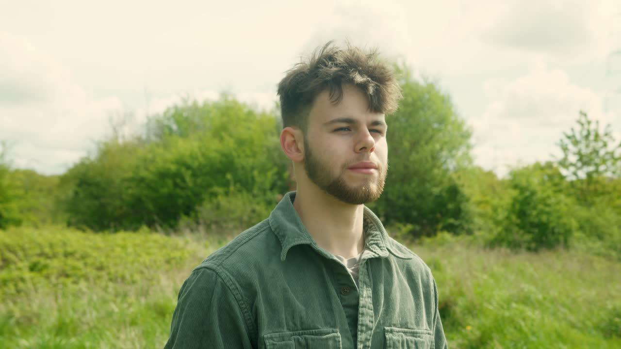 Man gazes outward in lush countryside under bright light, field and trees stretch behind him