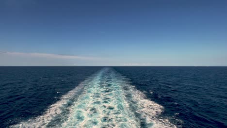 stern view of wide water wake left from cruise ship on sea water surface with horizon in background and shadow of people leaning over parapet of deck