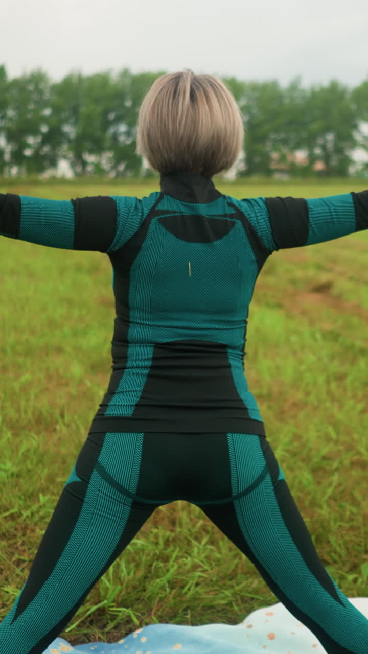 vista trasera de una mujer de pie en una alfombra de yoga, practicando la postura de triángulo de yoga con los brazos extendidos y las piernas separadas, en contra de un vasto paisaje de hierba con árboles y vegetación bajo un cielo nublado