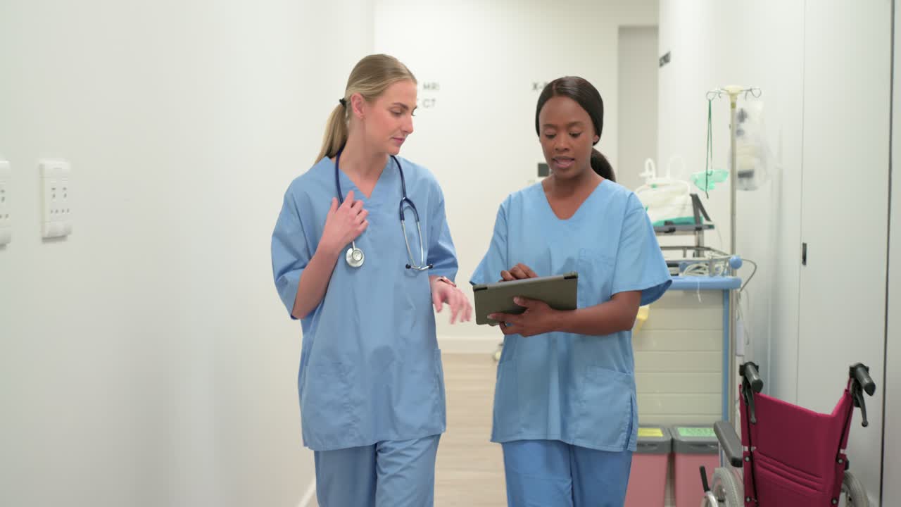Diverse female nurses in scrubs walking hospital hall checking tablet after imaging order, pointing