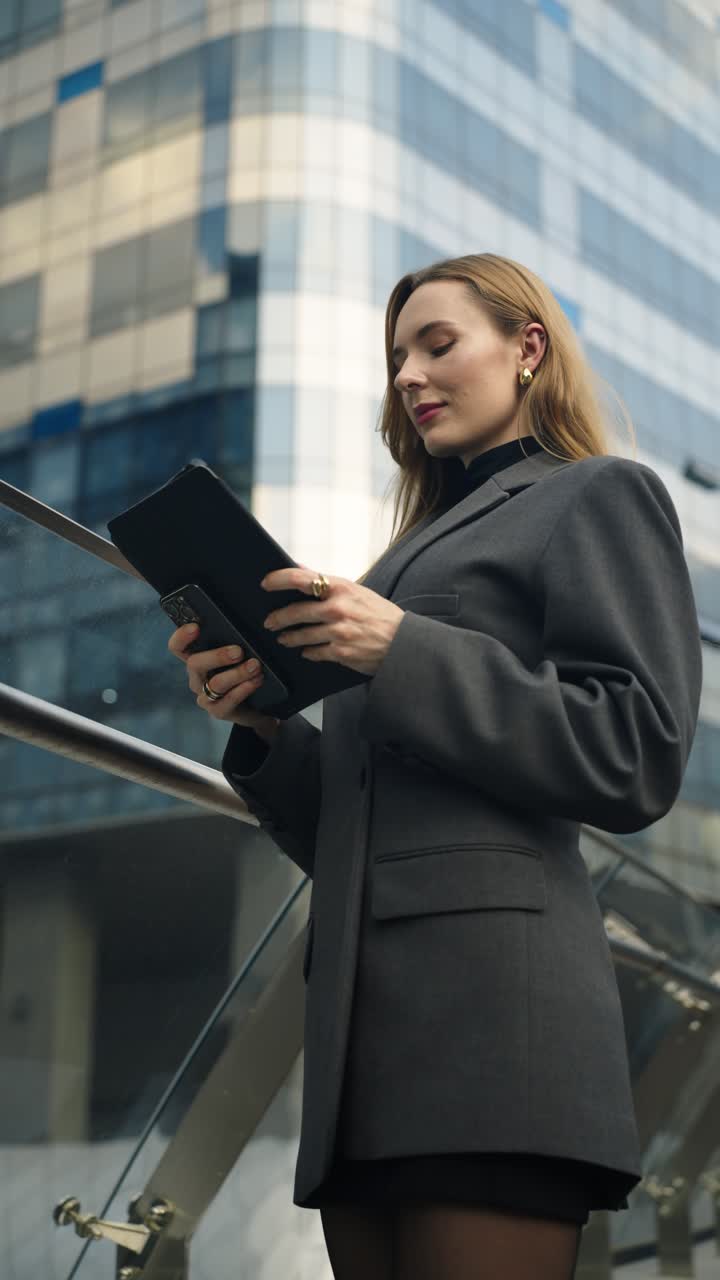Confident young woman in a blazer scrolls on a tablet and then calls on her phone outside a modern glass building - vertical, slow motion, parallax shot