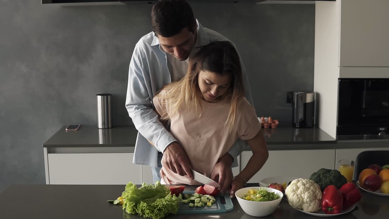 pareja amorosa en la cocina gris cocinando juntos. mujer joven triturando verduras junto con su marido. hombre alto de pie detrás y enseña a su esposa a cortar tomate. preparar el almuerzo de manera romántica