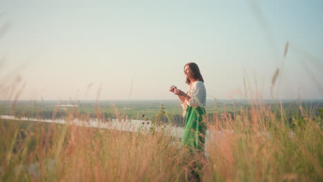 fair skinned woman in green dress standing on cliff blowing iridescent bubble above river valley at golden hour, tall grass swaying in gentle breeze creating peaceful outdoor