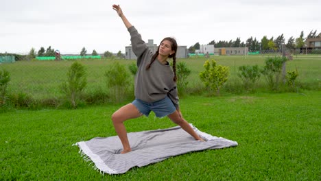 una mujer joven hace posturas de yoga guerrera en el césped en un día nublado y ventoso.