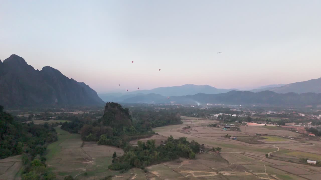 La luce del mattino si diffonde su Vang Vieng, Laos, mentre le mongolfiere si alzano sopra montagne calcaree, campi coltivati e valli nebbiose in un paesaggio pacifico circondato da serenità rurale e cime distanti.