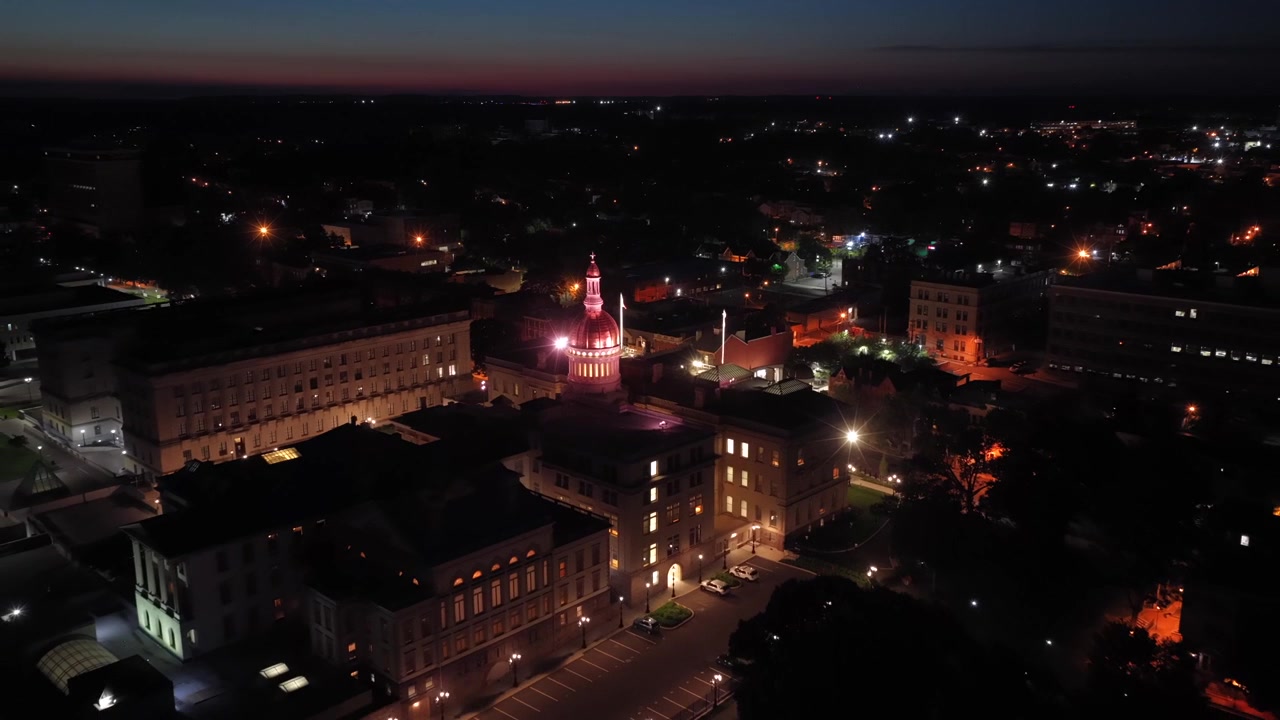 La cupola dorata illuminata del Campidoglio del New Jersey si erge sopra gli edifici del centro di Trenton di notte, con lampioni e luci della città che si diffondono sui tetti circostanti sottostanti. Ripresa aerea grandangolare all'indietro con drone