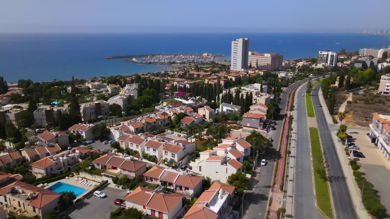 Drone shot of coastal marina area in Durres, Albania, featuring residential homes, tennis court, tall buildings, and clear blue sea under bright daylight with calm atmosphere
