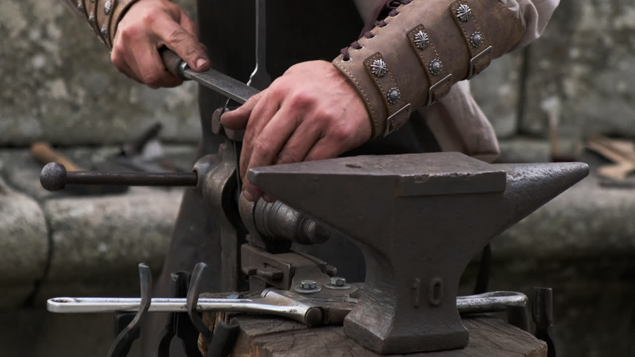 Static medium close-up as a blacksmith secures a forged piece in a bench vise to work and file it beside an anvil; precise shaping in a gritty workshop