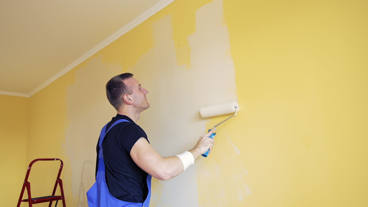 Room renovations at house. Paint being applied on a wall by a professional worker. Painter man painting the wall at home with paint roller and creamy color paint. Side view.