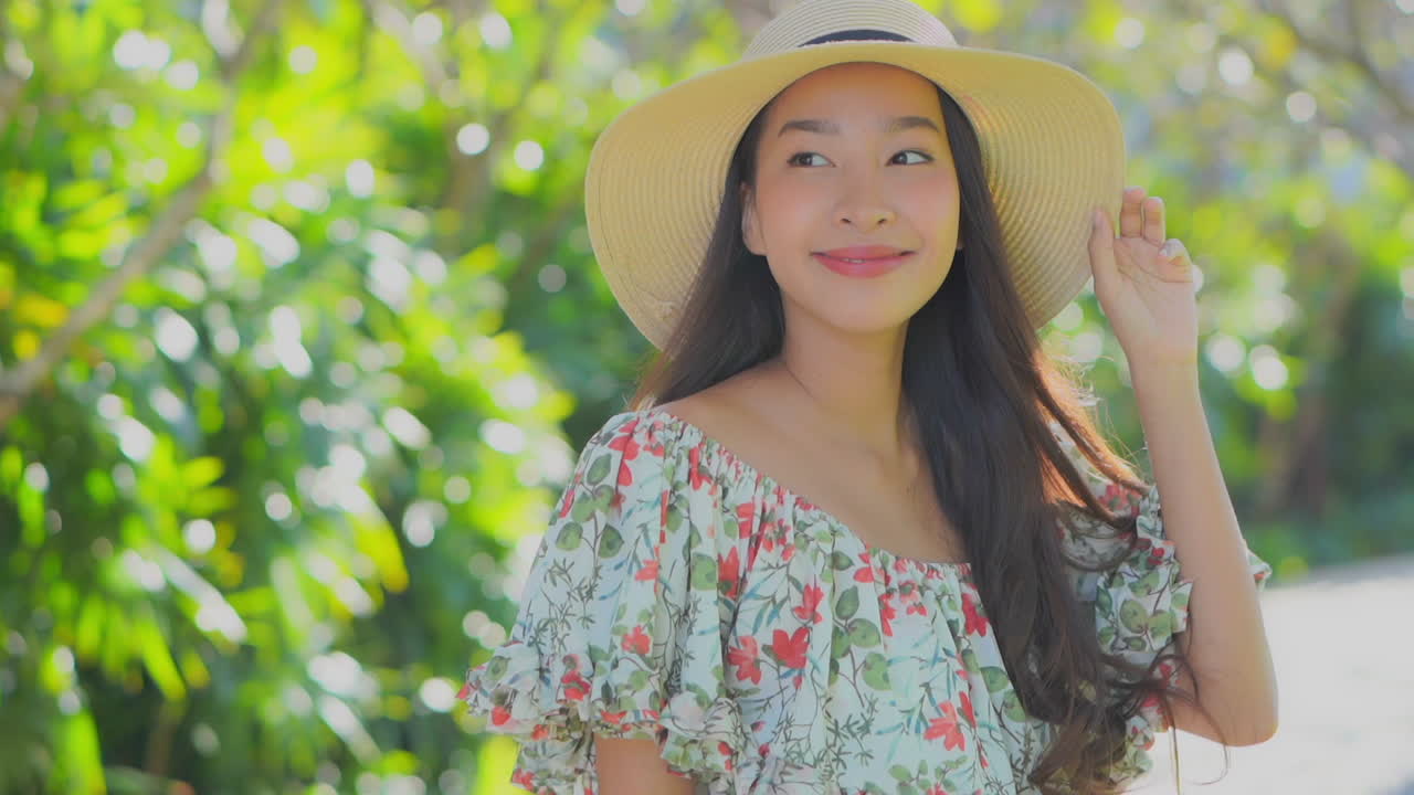 Close -up young, Asian, woman in a sun hat and flowered sundress makes eye contact then smiles and poses for the camera