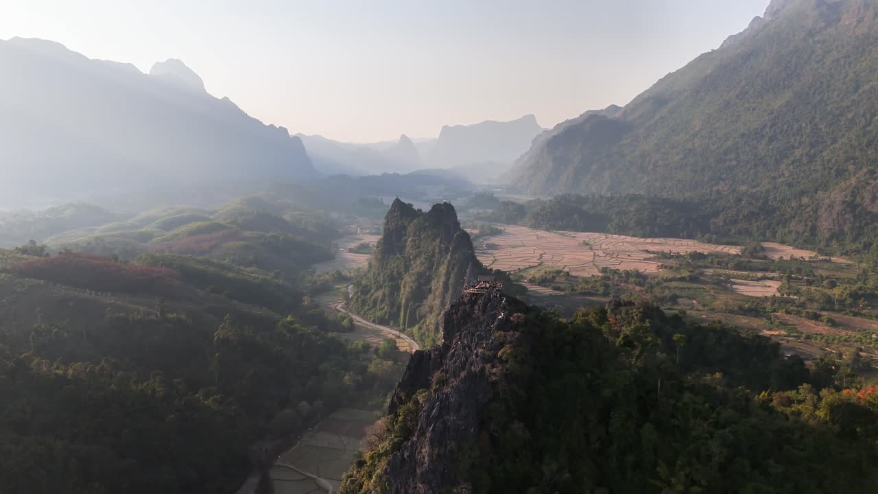 Ripide scogliere calcaree di Nam Xay si ergono sopra la valle rurale di Vang Vieng, Laos, con strade tortuose, risaie secche e colline boscose circostanti sotto un orizzonte montuoso morbido e nebbioso