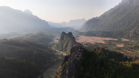 sharp limestone cliffs of nam xay tower above the rural valley of vang vieng, laos, with winding roads, dry rice fields, and surrounding forested hills under a soft, hazy mountainous horizon