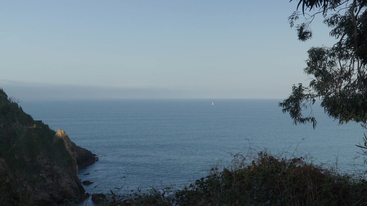 Scenic ocean view from coastal cliffs in Candás Asturias, Spain. Calm waters of the Cantabrian Sea stretch to the horizon with a small sailboat visible in the distance along northern Spain’s coastline