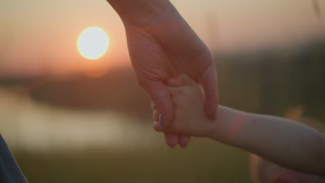 a close-up shot of a mother holding her younger son's hand, who is dressed in white, as they stand together watching a serene sunset over a tranquil lake. their faces are not visible