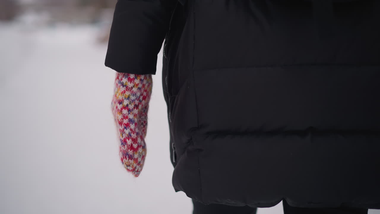 Female wearing black winter coat walking in snowy environment with colorful knitted mitten close up hand detail visible on side view cold frosty season blurred background outdoor urban lifestyle clothing warmth