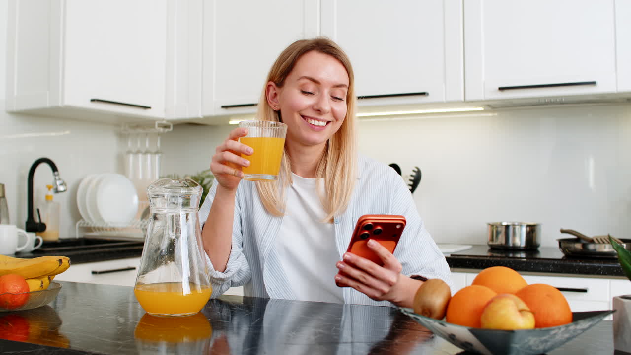 Joven bebiendo zumo de naranja natural durante su rutina de desayuno mientras lee noticias en su smartphone en la cocina de casa.
