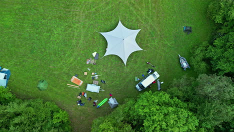 people camping on green meadow with huge tent and van, aerial top down view