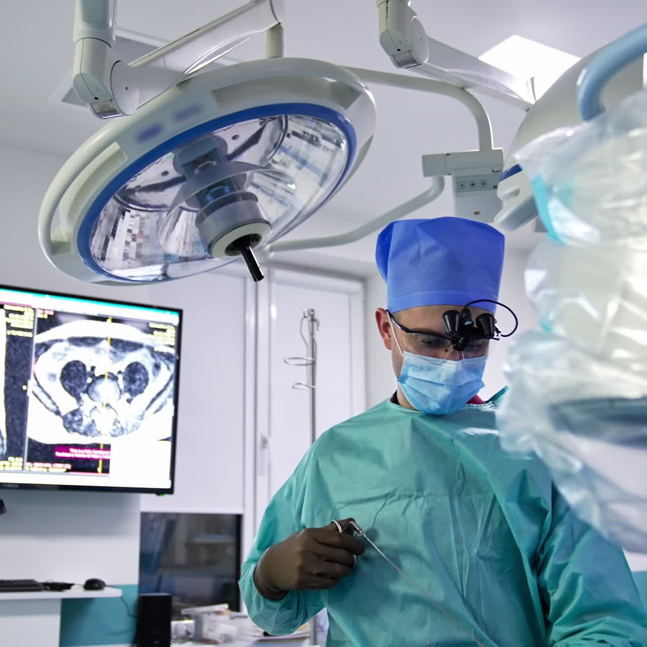 Neurosurgeon looks attentively in front of him and then pulls off the tool from a patient. Portrait of a doctor surrounded by surgery equipment and screen at the backdrop