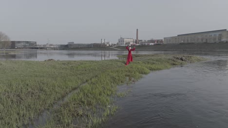 A-drone-shot-flying-away-from-a-model-with-red-dress-walking-on-a-small-island-with-a-city-in-a-background