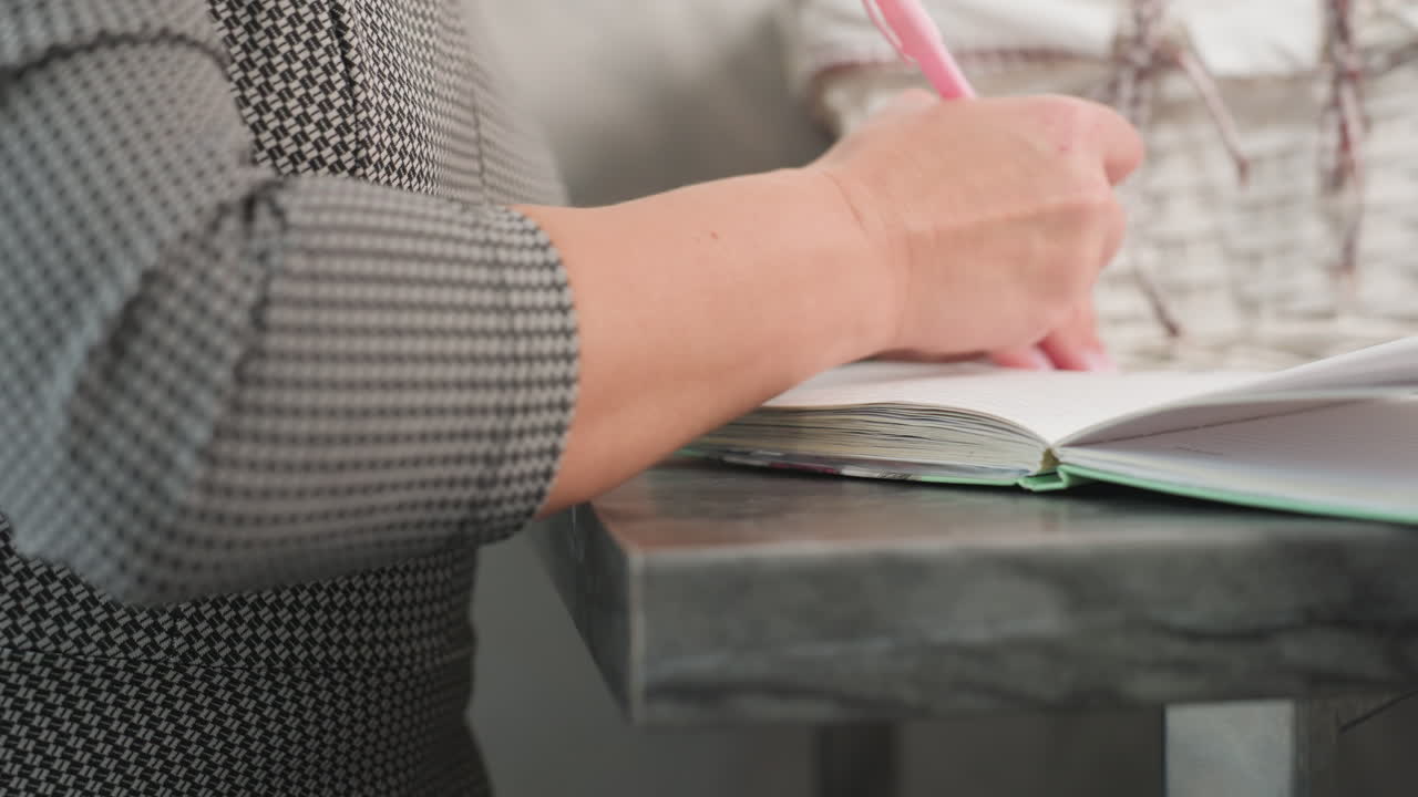 Close up of white lady flipping notebook page and writing with pink pen on marble table with blurred view of gift basket in background soft natural light