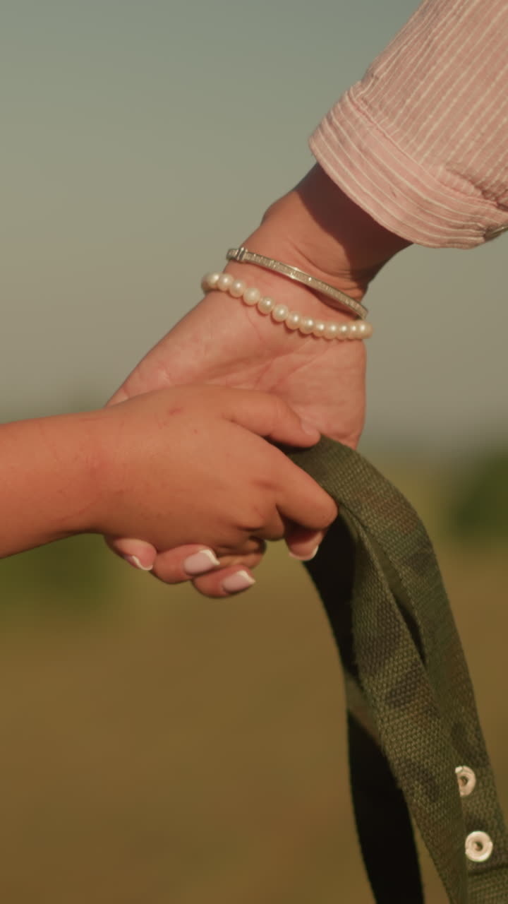 primer plano de un niño y un adulto tomados de la mano mientras agarran una cuerda juntos, un adulto lleva un brazalete de perlas, añadiendo un toque de elegancia contra el fondo de vastas tierras de cultivo abiertas bajo la cálida luz del sol